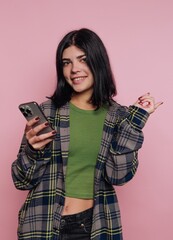Young woman in green top and plaid shirt smiles while using phone against pink background