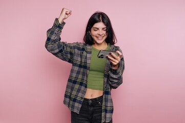 Young woman celebrates success while looking at her smartphone against a pink background