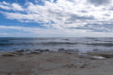 Low angle view of sandy shore with choppy waves, surfers on horizon, and distant landmass at Sagami Bay, Kamakura, Japan. Soft daylight and natural colors add a calm mood.