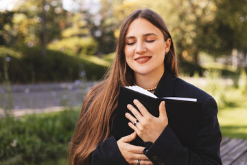 Pleased long hair brunette woman sitting on bench and reading book in park. Dreaming girl studying or read book for satisfaction. Enjoying girl. Woman hold her book diary hug near heart.
