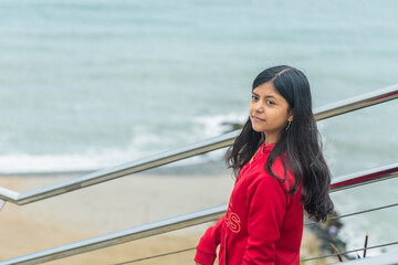 A girl wearing a red jacket stands on a railing overlooking the ocean