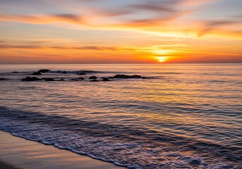A tranquil coastal sunrise paints the ocean in warm hues of orange and gold, showcasing gentle waves lapping at a sandy shore with dark rocks.