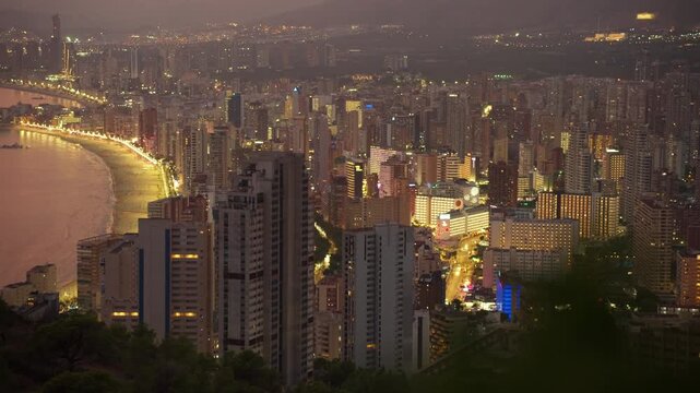 Panoramic aerial view of  spanish coastal city of benidorm at dusk, with glowing skyscrapers and city lights reflecting on the mediterranean sea, showcasing a vibrant night scene