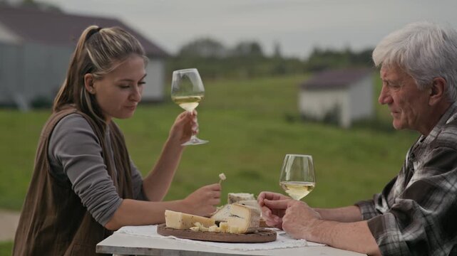 Side view shot of young Caucasian female farmer sitting at table with her senior father outdoors and discussing flavor of artisan cheese varieties made in their creamery