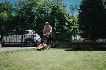 A man in a sleeveless shirt mows a green lawn in a sunny suburban yard. A decorative iron gate and a white car sit behind the fence, with trees and grass surrounding.