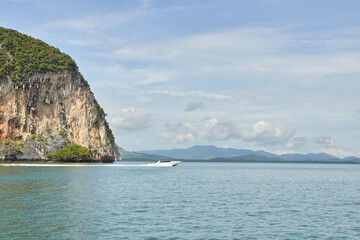 Fototapeta premium Speedboat navigating calm waters next to rocky cliffs under blue sky and cloudy horizon.