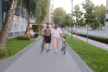 Naklejka premium A couple strolls down the walkway, each holding a bicycle. They are walking near a street.