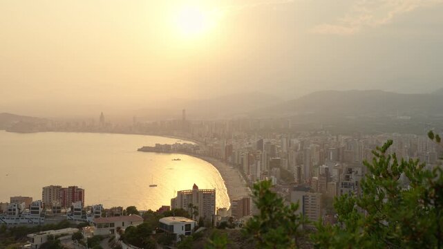 Panoramic elevated view of mediterranean coastal city of benidorm, spain, with sun setting over hazy sky, casting golden reflection on sea and famous levante beach