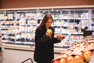 Young business long hair brunette woman girl buyer client consumer stands in shop store supermarket choosing food buying puts in basket. Woman choose fruits - apples. Healthy food.