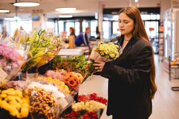 Long hair brunette woman wear jacket and choose bouquet of flowers, serious calm girl, she walk in supermarket, she is shopping after work. Garden market.