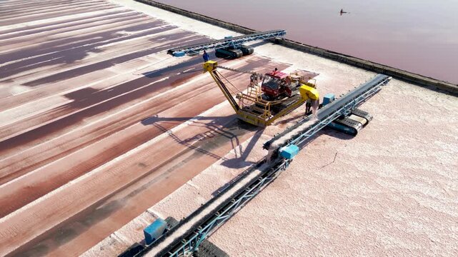 Drone top down aerial view of industrial salt harvesting equipment transferring freshly collected salt via conveyor system. Heavy machinery and workers operate at a salt production facility, showcasin