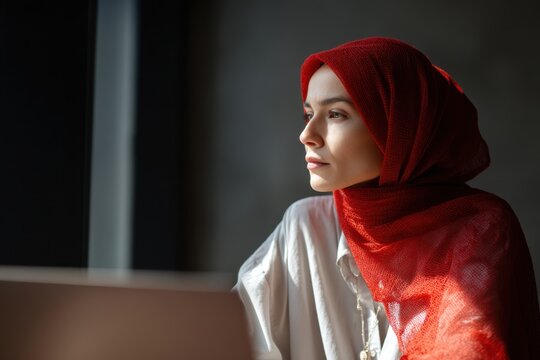 Woman in red hijab looking thoughtfully out a window in soft light