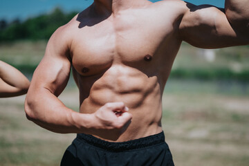 Obraz premium A man with a muscular physique confidently flexes his arms, showcasing strength and fitness under the sun, set against a nature-filled outdoor background.