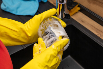 Fototapeta premium Hands wearing yellow gloves washing glass jar under running water in kitchen sink. Concept of cleaning, hygiene, sustainability, and eco-friendly home care.