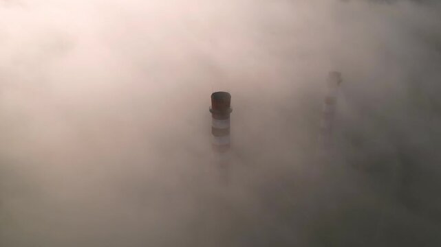 Aerial drone view of a tall orange and white industrial smokestack emerging from thick fog, with peeling paint, maintenance ladders, and a hazy pastel sky creating an atmospheric scene of aging infras