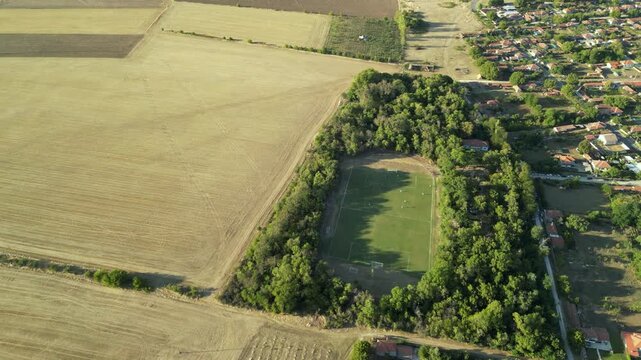 High-angle aerial drone view of a local football match on a green grass field, with players in yellow and white jerseys spreading out in midfield while long tree shadows stretch across the pitch in wa