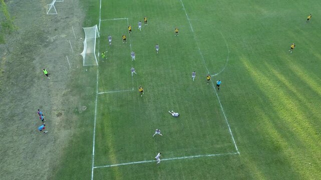 High-angle aerial drone view of a local football match on a green grass field, with players in yellow and white jerseys spreading out in midfield while long tree shadows stretch across the pitch in wa