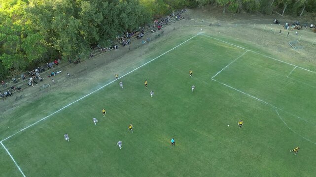 High-angle aerial drone view of a local football match on a green grass field, with players in yellow and white jerseys spreading out in midfield while long tree shadows stretch across the pitch in wa