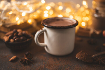 Hot chocolate in white enamel mug on dark surface with nuts, cocoa powder, spices, cupcake and festive lights, seasonal cozy drink and winter holiday concept