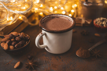 Hot chocolate in white enamel mug on dark surface with nuts, cocoa powder, spices, cupcake and festive lights, seasonal cozy drink and winter holiday concept