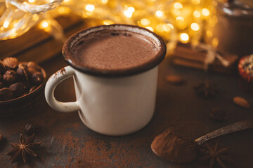 Hot chocolate in white enamel mug on dark surface with nuts, cocoa powder, spices, cupcake and festive lights, seasonal cozy drink and winter holiday concept