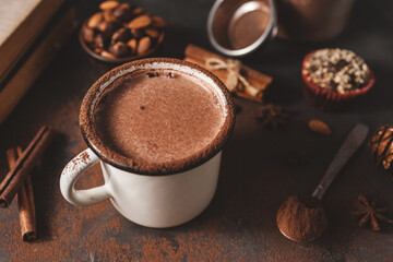 Hot chocolate in white enamel mug on dark rustic surface with nuts, cocoa powder, cinnamon sticks...