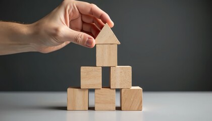 Hand builds house structure using wooden toy blocks. Triangle block placed on top of stacked square blocks. Image represents concepts like growth, foundation, development, planning, investment,