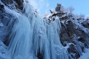 A breathtaking frozen waterfall flows down rugged cliffs, surrounded by snow-covered trees and rocks. The clear blue sky adds to the tranquil winter scene, inviting exploration
