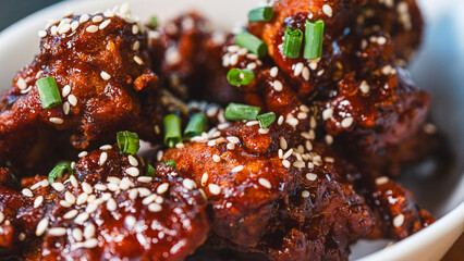 Sizzling, succulent Korean sticky chicken glistening with sauce, topped with sesame seeds and spring onions. A vibrant meal, beautifully presented in an elegant bowl within a stunning ambient setting.