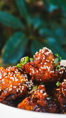 Sizzling, succulent Korean sticky chicken glistening with sauce, topped with sesame seeds and spring onions. A vibrant meal, beautifully presented in an elegant bowl within a stunning ambient setting.