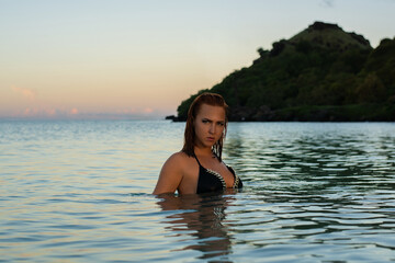 
Red-haired woman in a black bikini, sitting in shallow ocean water on a beach at sunset, with a dark, lush hill in the background.