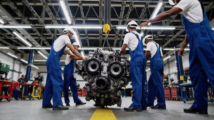 Professional mechanics wearing blue overalls, lifting heavy engine block with overhead crane, collaborating precisely inside spacious industrial garage workshop