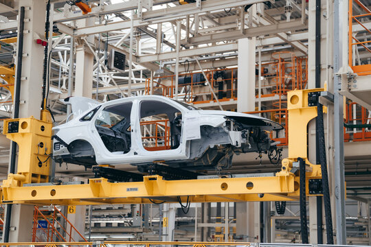 White car body is suspended on a yellow automated lift within a vast, multi-level automotive assembly plant