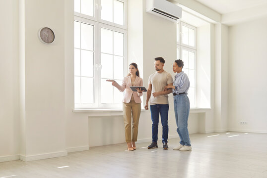 Realtor guides a young couple through a bright empty apartment with large windows and neutral interior. Female agent showing new house to clients. Move, real estate market and rent concept.  - Powered by Adobe