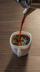Overhead view of black coffee being poured from a metal pot into a white ceramic cup, with bubbles forming on the surface on a wooden table in daylight with cast shadows.