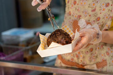 A chef wearing gloves holds a freshly made mochi or sweet dessert coated in dark crushed nuts in a paper container. The image captures the moment of serving street food