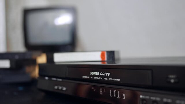 DVD player standing on the desk. Close up. VHS tape comes up and male hand takes it and puts on the pile.