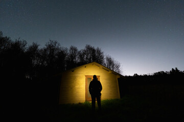 A mysterious silhouette of a person standing in the warm glow of a remote forest cabin under a starlit sky.