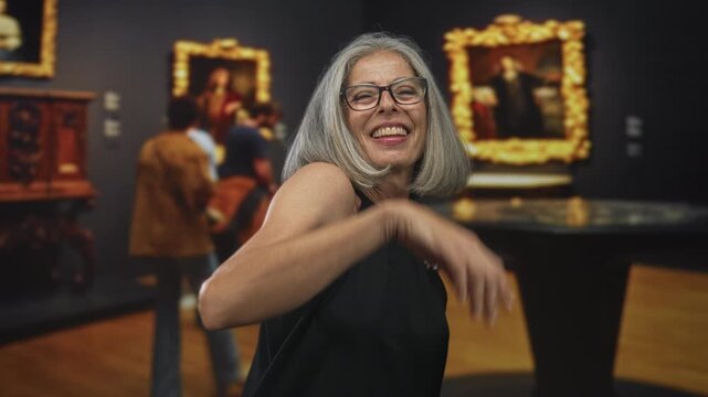 Woman dancing with raised arm and bare shoulder, wearing glasses and gray hair in a museum building; joyful carefree celebration.