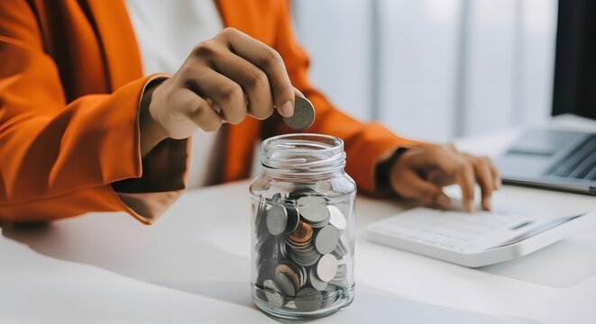 A person in an orange blazer is dropping a coin into a glass jar filled with coins, with a calculator and laptop in the background.