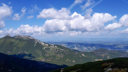 Fototapeta premium Scenic panoramic view of a mountainous landscape under a blue sky filled with fluffy clouds, showcasing lush greenery and distant valleys in summer sunlight.