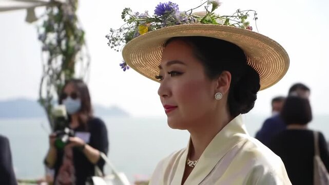 Elegant woman with flower hat at outdoor ceremony. Blurred people and sea in background, perfect for event photos