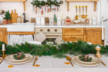 Elegant Christmas holiday table setting with greenery and simple decor on cozy kitchen background. Beautifully arranged holiday table with plates, candles and golden cultery on white tablecloth