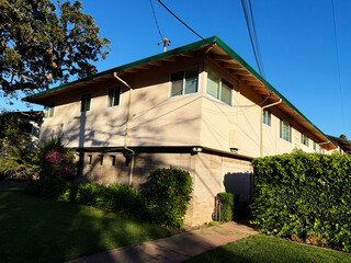Two-story low-rise beige apartment building with green roof, multiple windows, and landscaped garden with hedges and flowering plants under clear blue sky in a suburban residential area