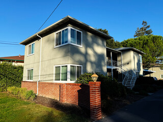 Two-story residential low-rise apartment building with stucco exterior, red brick base, white framed windows, and outdoor staircase surrounded by green hedges and trees