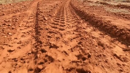 A low-angle, dynamic shot captures a rugged, dusty dirt road winding through a natural, arid landscape. Deep tire tracks are clearly visible in the reddish-brown earth, revealing a challenging and unp - Powered by Adobe