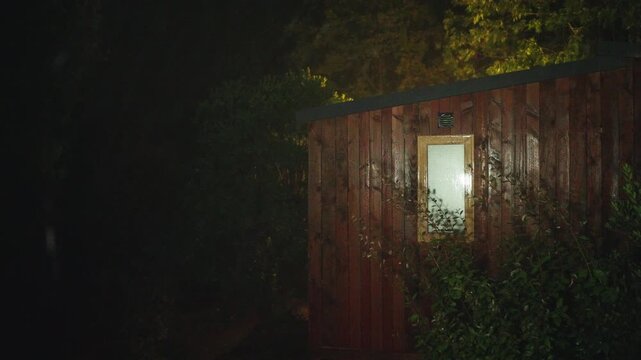 Dark rainy night scene showing a wooden cabin exterior illuminated by artificial light with visible rainfall and surrounding plants