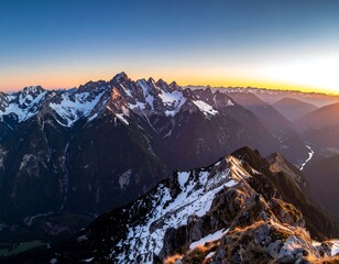 An aerial view of a snow-capped mountain range at dawn, with jagged peaks and valleys. The sun sets along the horizon