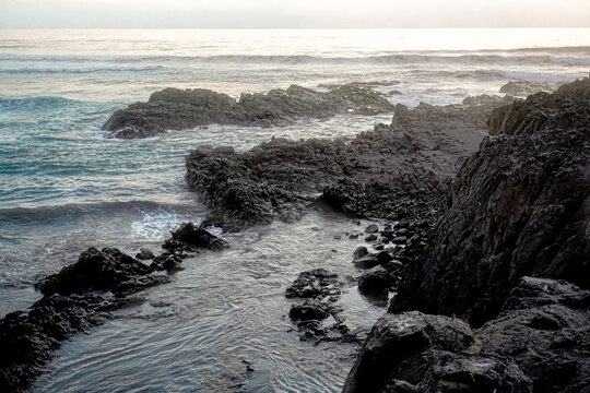 Dram&aacute;tico paisaje costero con poderosas olas rompiendo contra rocas escarpadas al atardecer."