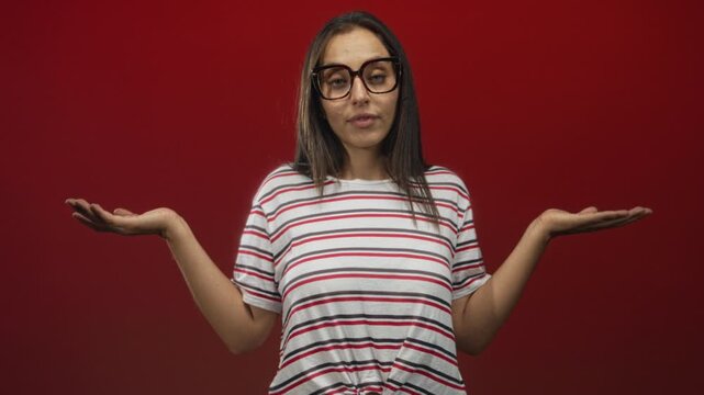 Woman with glasses in striped shirt holding both palms up in red studio with closed eyes and neutral expression; uncertainty.